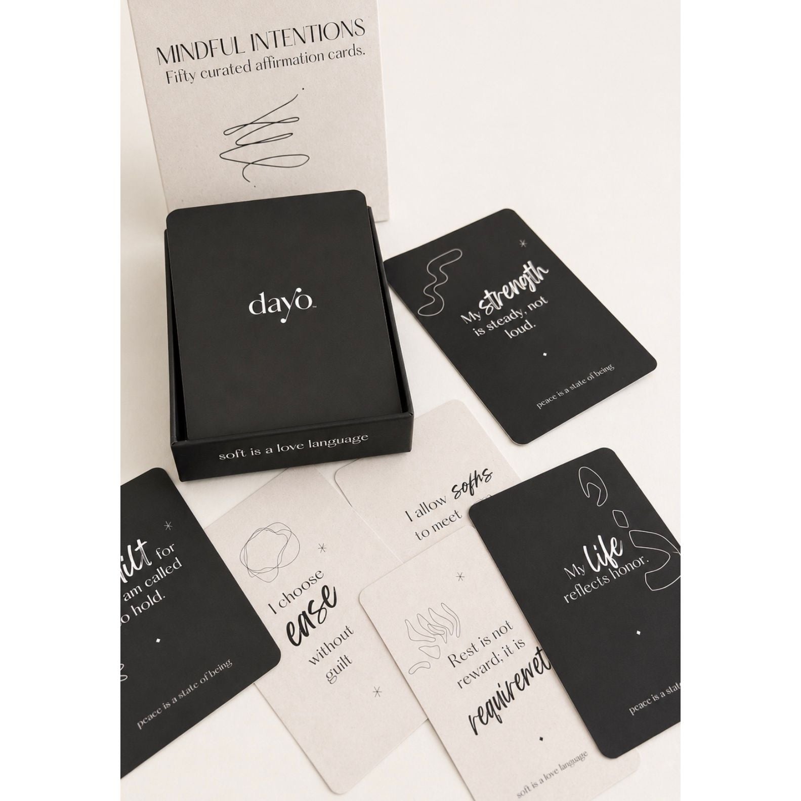A boxed card deck with several black and off-white affirmation cards arranged on a white background. The box lid shows the brand name 'clay6' and the top card reads 'MINDFUL INTENIONS FIFTY curated affirmation cards.